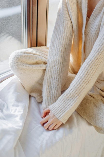 Person wearing a beige knitted sweater and pants sitting on a bed.